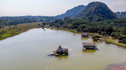 Scenic aerial view of a tranquil lake with rustic huts surrounded by lush greenery and hills under a clear sky.