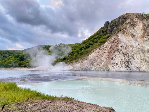 Scenic view of a steaming hot spring surrounded by lush greenery and rocky hills under a cloudy sky.