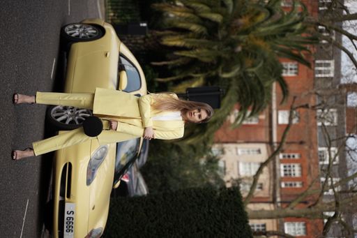 A Caucasian female in a white and yellow outfit posing for the camera outdoors in front of the car