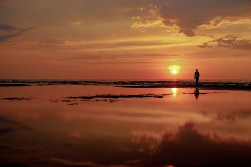 A solitary figure walks along the shore during a vibrant sunset, reflecting on the calm water.
