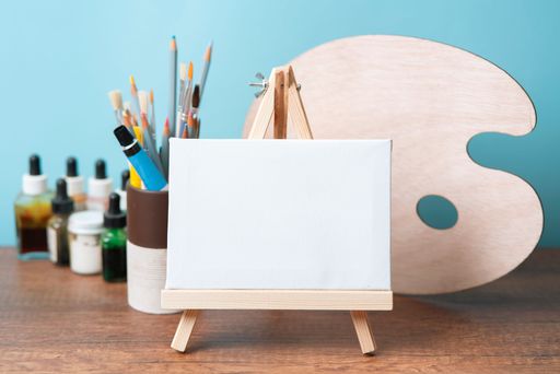 Front view of small wooden easel with blank canvas on wooden table decorated with glass with paint tubes, brushes and palette against blue background