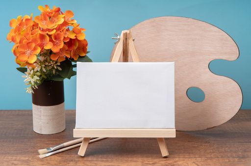 Front view of small wooden easel with blank canvas on wooden table decorated with glass with flowers, paintbrushes and palette against blue background