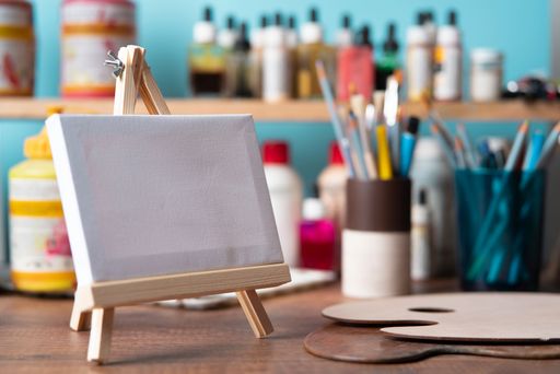 Front view of small wooden easel with blank canvas on wooden table against background of shelves with paint tubes, brushes and pencils