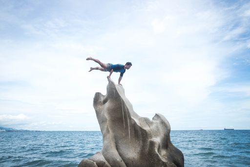 An athletic Hispanic man doing a horizontal handstand on a rock against an ocean view