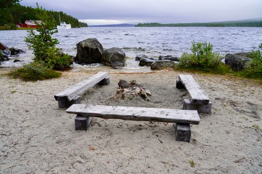 Wooden benches around a campfire pit on a sandy lakeshore with lush greenery and distant boats, under a cloudy sky.