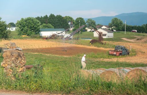A rural farm scene with an elderly man working in a field, surrounded by hay bales, old truck, and distant mountains under a clear sky.