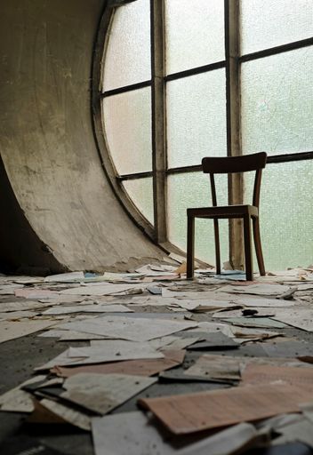 A vertica shot of a chair in front of a round window with papers and book pages lying on the ground