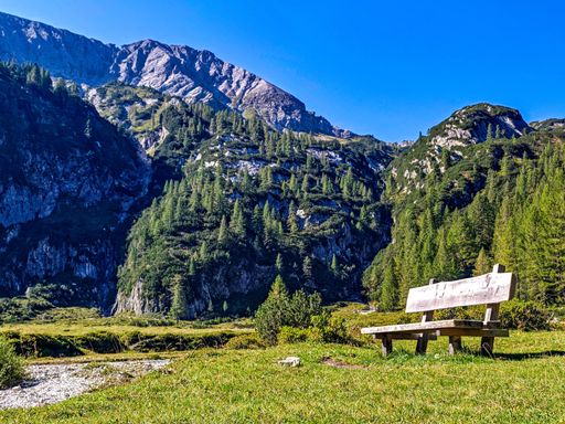 A serene mountain landscape featuring a wooden bench under a clear blue sky, surrounded by lush greenery and rocky peaks.