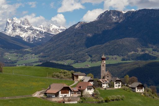 Scenic view of a picturesque village with a church tower, nestled in lush green hills, with snow-capped mountains in the background.