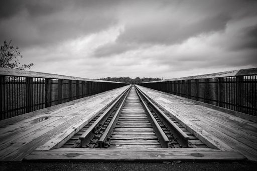 Black and white image of a wooden railroad bridge stretching into the distance under a cloudy sky.