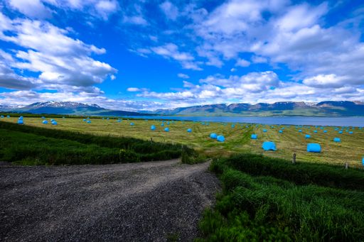 A road near a grassy field with rolled up hays in a blue plastic bags and mountains in the distance under a cloudy sky