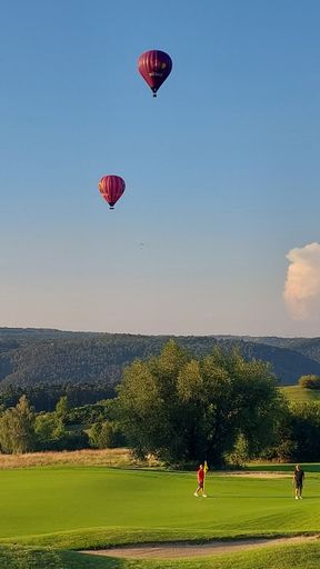 Hot air balloons soar over a lush green golf course with players enjoying a sunny day, set against a backdrop of rolling hills and clear blue sky.