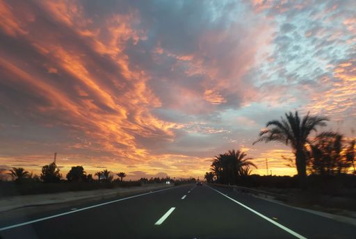 A scenic highway at sunset with dramatic clouds and palm trees on the horizon.