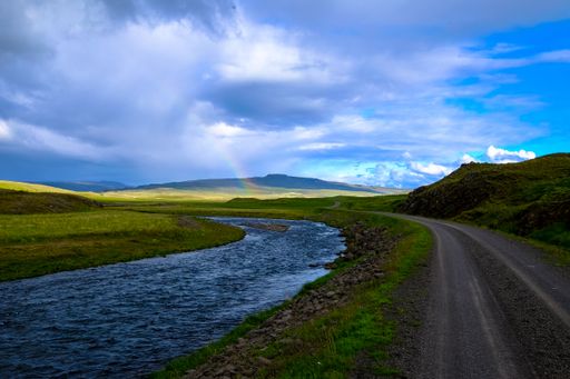 A river in the middle of a road and grassy field with a rainbow in the distance at daytime