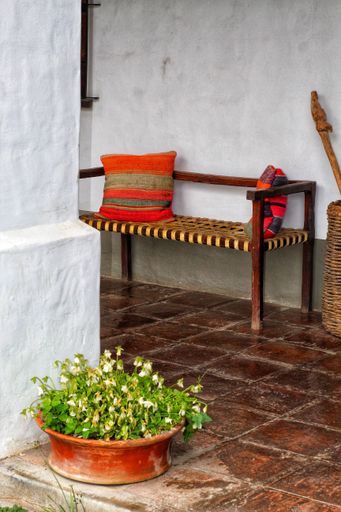 Rustic outdoor seating area with a wooden bench and colorful cushions, next to a potted plant on a tiled floor.
