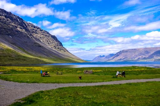 A beautiful shot of a pathway in the middle of a grassy field with people sitting on a bench and mountains in the distance under a cloudy sky