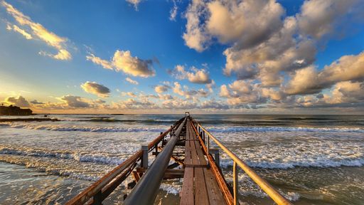 A scenic view of a pier extending into the ocean under a vibrant sky with fluffy clouds at sunset.