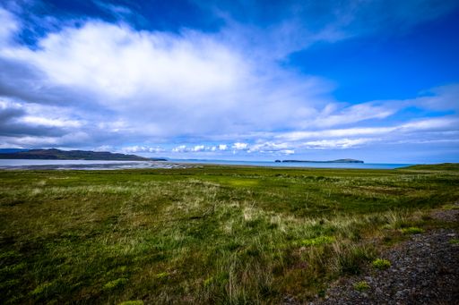 A grassy field near the sea with mountains in the distance under a cloudy sky