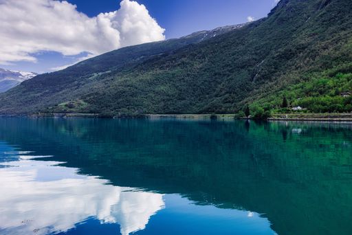 Serene lake with lush green mountains and clouds reflected in clear water, under a bright blue sky.