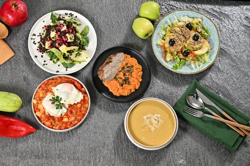 A top view of a variety of healthy dishes including salads, soup, and eggs on a grey stone surface with fresh fruits and vegetables.