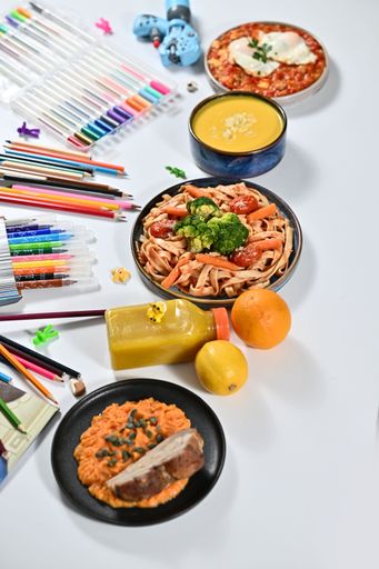 Colorful assortment of food and art supplies on a white table, featuring pasta, soup, pizza, and a variety of pencils and markers.