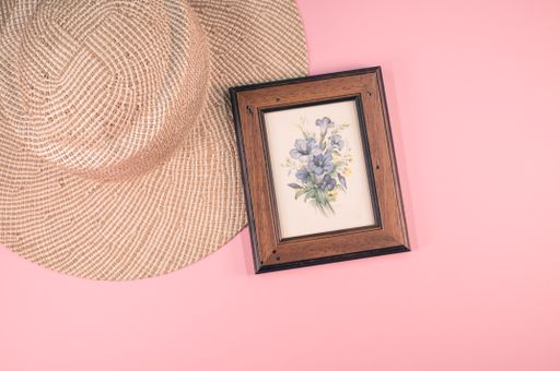 A top view of a framed picture of flowers with a sun hat on a pink surface