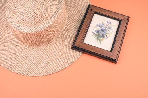 A closeup of a straw hat and blue flowers painting with a wooden frame, on an orange surface