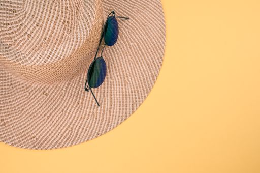 A top view of a sun hat with a pair of sunglasses on it on a yellow surface with space for text