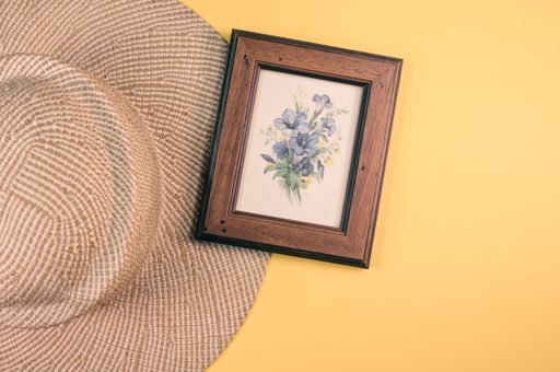 A top view of a framed picture of flowers with a sun hat on a yellowsurface