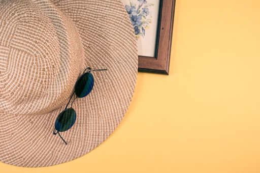 A closeup of a straw hat, blue flowers painting with a wooden frame, and sunglasses on a yellow surface