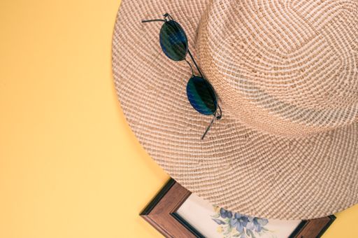 A top view of a straw hat, sunglasses, and blue flowers painting with a wooden frame, on a yellow surface