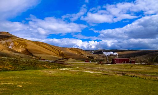 A dry grassy field with a red factory in the distance near the mountains under a cloudy sky