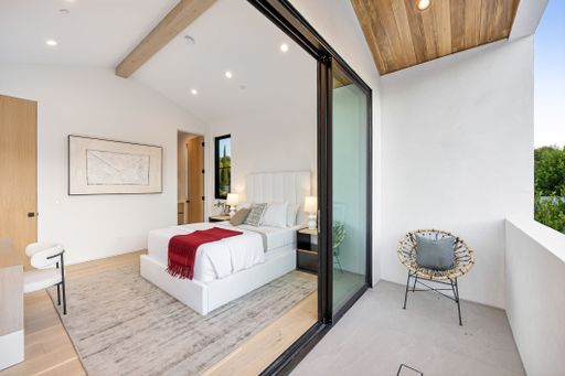 Modern bedroom with elegant decor, featuring a cozy bed with a red throw, large art piece, and a balcony with a chic chair.