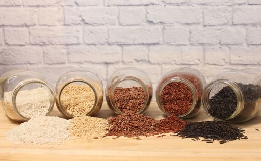 Variety of rice grains in glass jars on a wooden table against a brick wall background.