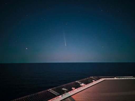 A serene night sky over the ocean with a visible comet and stars, captured from a ship's deck.