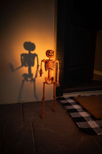 A Halloween-themed skeleton decoration with a pumpkin head casting a shadow on a wall near a doorstep.