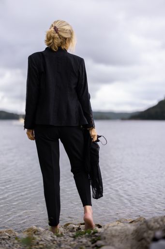 A vertical shot of a blonde woman in a suit standing at the shore, admiring the sea under a cloudy gray sky