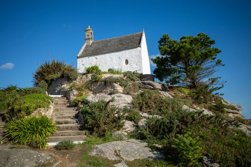 Charming white chapel on a rocky hill with lush greenery under a clear blue sky, capturing tranquility and architectural beauty.