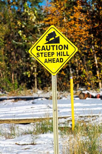 A vertical of a metal yellow sign with " CAUTION STEEP HILL AHEAD" written on it on the roadside