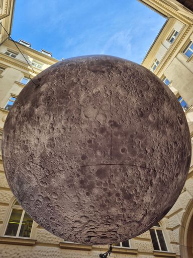 A large moon sculpture suspended in a courtyard under a clear blue sky, surrounded by historic buildings.
