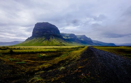 A road in the middle of grassy fields and mountains in the distance under a cloudy sky