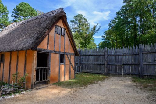 Rustic thatched-roof cottage with wooden fence in a lush forest setting under a clear blue sky.