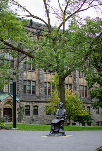 A statue of a seated figure in front of a historic stone building surrounded by lush green trees, capturing a serene campus scene.