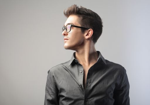 A closeup of an attractive Caucasian male wearing glasses and a formal shirt in front of a gray wall