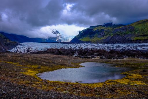 A wide shot of a small pond surrounded by green mountains and white rock formations under a cloudy sky