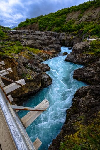 A vertical shot of a river flowing in the middle of cliffs and grassy hills under a cloudy sky