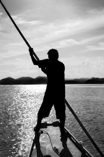 A grayscale shot of a male standing on the boat on the shiny water under a cloudy sky