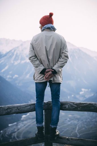 A vertical shot of a man wearing a red hat and standing on a wooden fence with mountains in the background