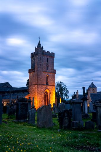 Historic church tower illuminated at dusk, surrounded by ancient gravestones under a dramatic cloudy sky in a serene cemetery setting.