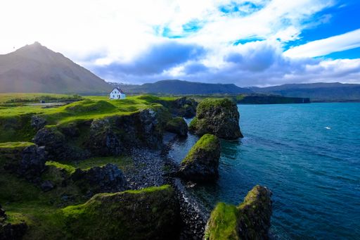 An aerial shot of mossy rocks near the water with a white house in the distance near a mountain under a cloudy sky